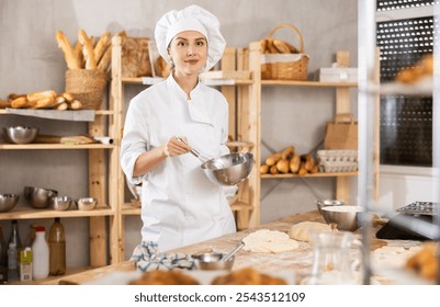 Professional young female baker in white uniform using whisk to mix ingredients in metal bowl, standing in small artisan bakery, surrounded by variety of freshly baked bread and tools - Powered by Shutterstock