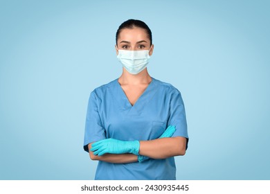 Professional young female nurse in blue scrubs, surgical mask, and gloves standing with arms crossed, ready for medical duties on tranquil blue background - Powered by Shutterstock