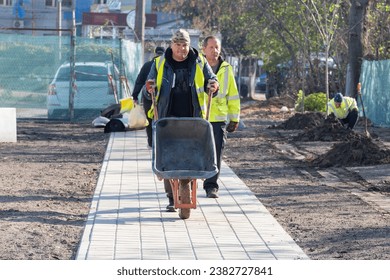 A professional young man-gardeners pushing wheelbarrow along the walkway in the city park or square near newly planted trees at spring or autumn. - Powered by Shutterstock