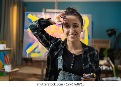 Professional young woman artist rubs her face with hand from paint pa three on camera with smile. Authentic creative studio with large canvas and abstract painting