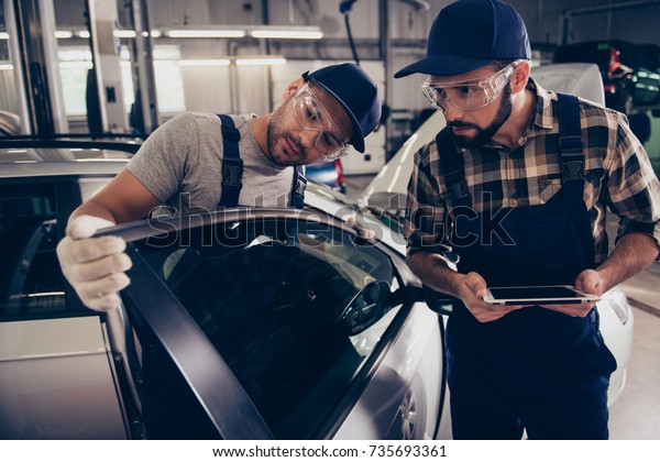 Professionalism, maintenance. Two busy expert\
engineers at mechanical work shop, in special safety outfit\
uniform, protective spectacles, focused, serious, concentrated,\
estimating upgrading of\
car