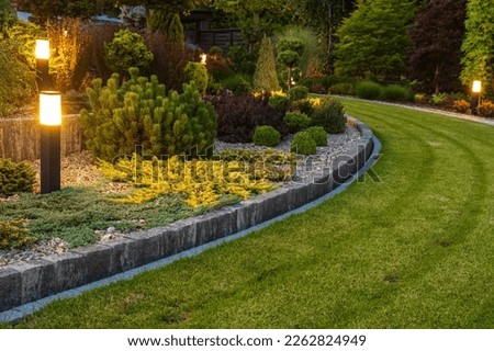 Professionally Landscaped Backyard Garden with Evenly Mowed Lawn and Trimmed Shrubs Illuminated with Outdoor Bollard Lamps.