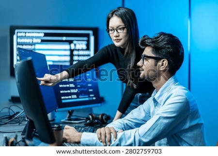 IT professionals collaborating in a dark server room. Female data analyst pointing at computer screen discussing software code with male programmer. Cyber security team monitoring digital network.