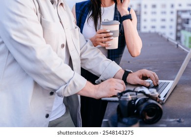 Professionals collaborating on laptop, enjoying coffee on urban rooftop. Male typing on keyboard, female holding coffee cup, teamwork and creativity. Camera in foreground, photography work environment - Powered by Shutterstock