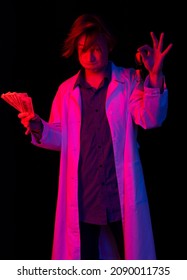  Professor Conducting Experiments. Guy In A Dressing Gown Holds Glasses And Money. Colored, Blue-red Background. Blur