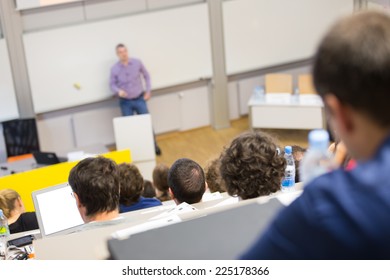 Professor Giving Presentation In Lecture Hall At University. Participants Listening To Lecture And Making Notes.