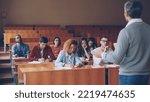 Professor is reading lecture to group of multiracial students standing in classroom with papers and speaking while young people are listening to him and writing sitting at desks.
