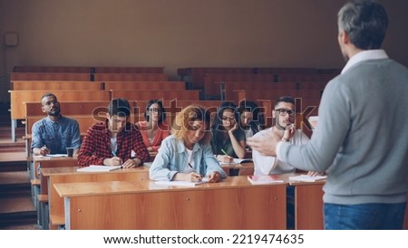 Professor is reading lecture to group of multiracial students standing in classroom with papers and speaking while young people are listening to him and writing sitting at desks.