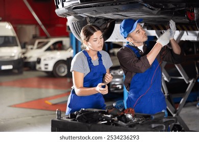 Proficient car mechanic checking operability of auto-element while girl in blue overalls watching him attentively in auto-repair shop