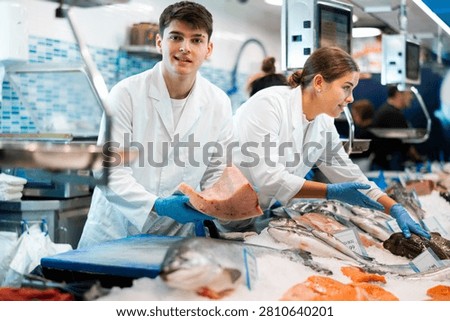 Proficient shop assistant in white work clothes holding in hands tuna in fish shop with large stock