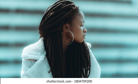 Profile of an adorable young black female with big dreadlocks, round earrings, and piercing in her nose, she is looking aside while standing outdoors with a striped facade in defocused background