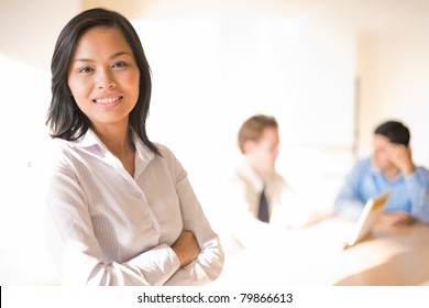 Profile of beautiful Asian businesswoman standing, smiling, looking at camera in white blouse, arms crossed in front foreground focus and male work colleagues in background. High key horizontal