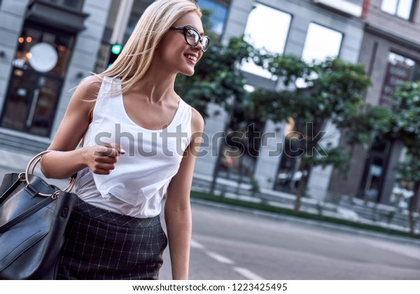 Profile of a beautiful\
businesswoman with glasses and handbag. She crosses the road and\
looks for cars nearby