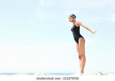 Profile Body View Of A Beautiful Young Woman Swimmer Diver Preparing To Dive Off A Rocky Cliff By The Sea, Wearing Goggles Against A Sunny Blue Sky. Health And Beauty, Sport Lifestyle.