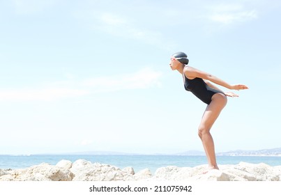 Profile Body View Of A Beautiful Young Woman Swimmer Diver Preparing To Dive Off A Rocky Cliff By The Sea, Wearing Goggles Against A Sunny Blue Sky. Health And Beauty, Sport Lifestyle.