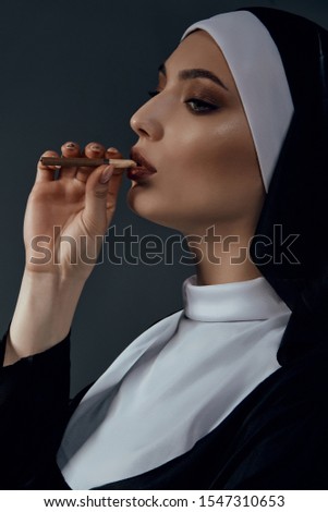 Profile close-up portrait of a nun, posing on a black background. She wearing dark nun's clothing. The nun brings a cigarette to her mouth and looking forward. 