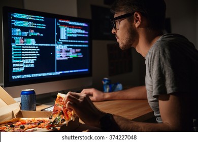 Profile of concentrated young software developer eating pizza and coding at home