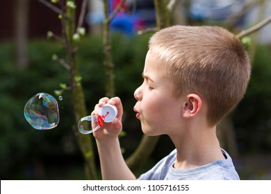 Profile Of Cute Handsome Small Blond Child Boy With Funny Serious Expression Blowing Colorful Transparent Soap Bubbles Outdoors On Blurred Green Summer Background. Joy Of Careless Childhood Concept.