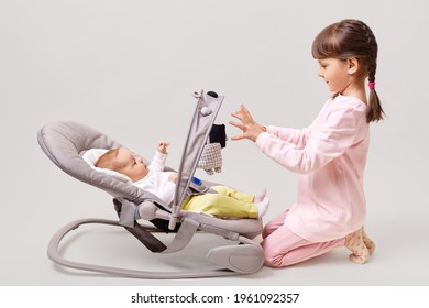 Profile Of Cute Little Girl With Pigtails Wearing Pink Casual Clothing Playing With New Born Baby Sister Girl Lying In Bouncer Chair Play, Isolated Over White Background.