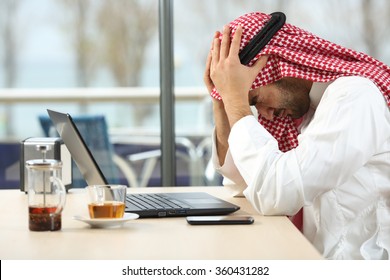 Profile Of A Desperate And Alone Arab Saudi Man With A Laptop Online In A Coffee Shop With A Window In The Background. Bankruptcy Concept