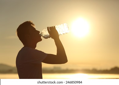 Profile of a fitness man silhouette drinking water from a bottle at sunset with the sun in the background