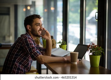 Profile Of Handsome Man Working In Restaurant. Happy Man Talking Over Mobile Or Smart Phone, Looking At Window While Sitting In Front Of Laptop Computer.