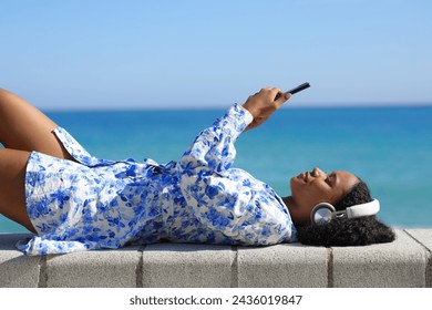 Profile of a happy black woman wearing headphone watching media on phone lying on the beach - Powered by Shutterstock