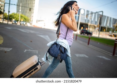 Profile Of Happy Woman Pulling Rolling Suitcase And Carrying Handbag While Talking On Mobile Phone Outdoors In City