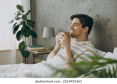 Profile happy young man wear casual clothes t-shirt pajama lying in bed drink coffee tea hold cup close eyes rest relax spend time in bedroom home in own room hotel wake up dream be in good mood day - Powered by Shutterstock