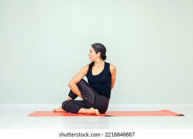 Profile Of Hispanic Mature Female In Sportswear Practicing Yoga Stretches , Sitting In Ardha Matsyendrasana Exercise, Variation Of Half Lord Of The Fishes Pose. Black Pants And Top, Studio Background.