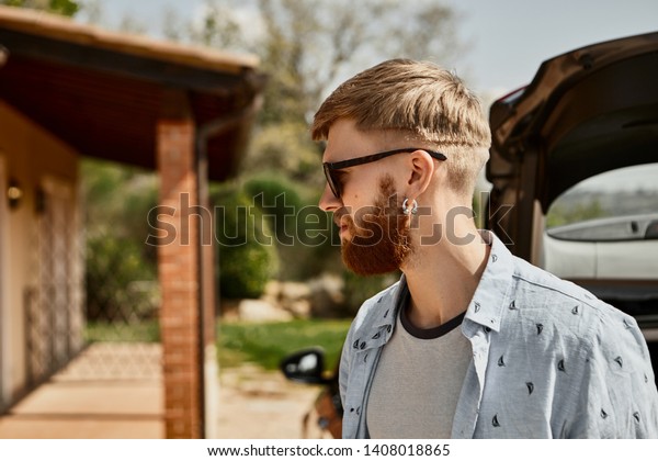 Profile image of fashionable young hipster guy
with trendy hairstyle and thick ginger beard going home from nice
walk on sunny summer day, wearing stylish rectangular sunglasses
and earrings