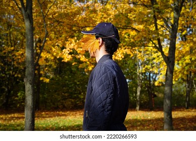 Profile of a man with an autumn leaf on his face. A young man in a blue jacket and cap, with black curly hair stands sideways in the middle of the frame, his face covered by an orange maple leaf. - Shutterstock ID 2220266593