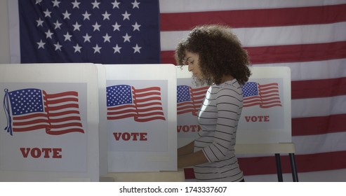 Profile, medium shot, young Hispanic woman in polling station, voting in a booth with US flag in background.