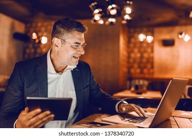 Profile of middle aged businessman dressed smart casual typing on laptop and holding tablet while sitting in cafeteria. - Powered by Shutterstock