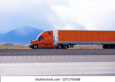 Profile of orange bonnet big rig pro semi truck with long dry van orange semi trailer running on the flat road with blue mountain on background in Utah for long haul commercial cargo delivery trip - Shutterstock ID 1071793688