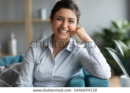 Photo of Profile picture of smiling beautiful millennial Indian girl sit rest on couch in living room, portrait of happy young ethnic woman relax on comfortable sofa at home look at camera feel positive