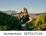 A profile picture of a young beautiful woman with long straight blonde hair, wearing black t-shirt, drinking from yellow thermo bottle, gorgeous landscape of mountains covered with coniferous forest