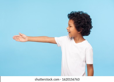 Profile Of Polite Cheerful Preschool Boy With Curly Hair In White T-shirt Giving Hand To Handshake, Gullible Trusting Child Meeting With Friendly Smile. Indoor Studio Shot Isolated On Blue Background