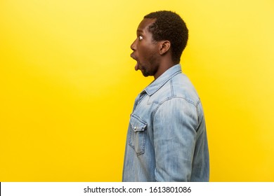 Profile Portrait Of Amazed Young Man With Bristle In Denim Casual Shirt Looking Left Side With Open Mouth, Astonished Expression, Blank Copy Space For Text. Studio Shot Isolated On Yellow Background