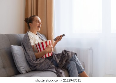 Profile portrait of of attractive impressed woman with bun hairstyle sitting on couch wrapped in blanket and watching film with tasty snack, enjoying interesting film or tv show.