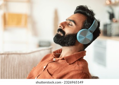 Profile portrait of bearded arab man in wireless headphones reclining on sofa, resting with eyes closed at home. Relaxed guy listening to peaceful music in headset on weekend, copy space - Powered by Shutterstock