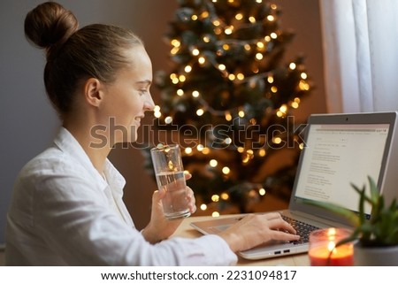Profile portrait of beautiful woman with bun hairstyle wearing white shirt, working on pc computer and holding glass in hands, drinking water, with xmas tree on background.