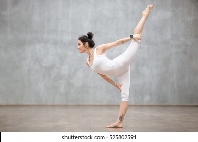 Profile portrait of beautiful young woman wearing white sportswear working out against grey wall, doing yoga or pilates exercise. Standing in variation of Natarajasana, Lord of the Dance. Full length