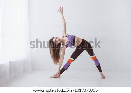 Profile portrait of beautiful young woman working out against white wall, doing yoga or pilates exercise. Standing in Utthita Trikonasana, extended triangle pose.