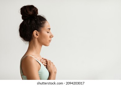 Profile Portrait Of Calm Young Woman With Hands On Chest And Closed Eyes, Side View Shot Of Peaceful Millennial Female Meditating Or Praying While Standing Over White Background Indoors, Copy Space