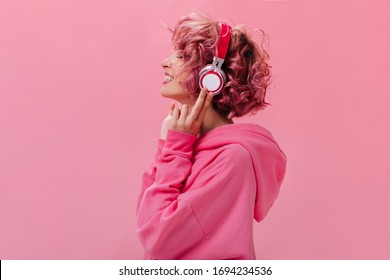 Profile portrait of charming pink-haired woman in massive headphones listening to music on isolated background. Curly girl in hoodie smiles. - Shutterstock ID 1694234536