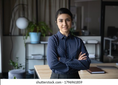 Profile portrait of confident young indian businesswoman standing in modern office with folded hands. Professional financial consultant lawyer ceo manager startup founder looking at camera.