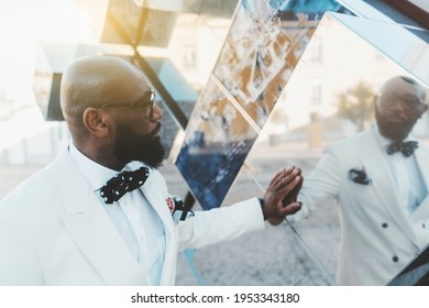 A profile portrait of a dapper handsome bald bearded mature black man in a white costume with a dotted bow tie, touching the surface of a mirror cube outdoors and looking at the reflection of himself
