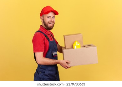 Profile Portrait Of Delivery Man Wearing Blue Uniform And Red Cap Holding Cardboard Boxes And Paper House Safe Door-to-door Delivery. Indoor Studio Shot Isolated On Yellow Background.