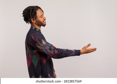 Profile Portrait Friendly African Man With Dreadlocks Stretching Hand Welcoming Guests With Toothy Smile On Face, Nice To Meet You. Indoor Studio Shot Isolated On Gray Background